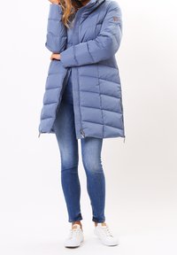 Ljust blå pufferjacka med dragkedja fram, huva och quiltat tyg. Bärs över blå jeans och vita sneakers, med sidodragkedjedetaljer.