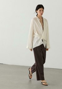 Vrouw loopt binnenshuis, draagt een loszittende witte blouse met lange mouwen, donkere brede broekspijpen en zwarte teenslippers tegen een effen achtergrond.