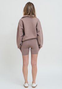 Felpa marrone oversize con spalle scese, abbinata a pantaloni corti marroni aderenti. L'outfit presenta una texture liscia e un design minimal.