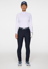 Kvinna i vit långärmad skjorta, marinblå byxor med bälte, blå och vita sneakers, marinblå keps och vita handskar, stående mot en helt vit bakgrund.