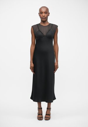 Vestido negro sin mangas con corsé de malla transparente, tela suave y una silueta fluida. Combinado con sandalias negras con tiras.