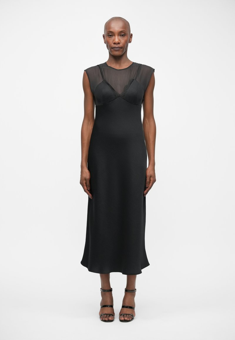 Μαύρο αμάνικο φόρεμα με διάφανη διαφάνεια στο σώμα, λεία ύφανση και αέρινη σιλουέτα. Συνδυάζεται με μαύρες σανδάλες με λουράκια.