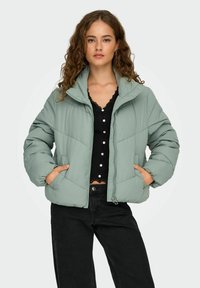 Lys grøn pufferjakke med høj krave, lynlåslukning foran og sidelommer. Struktureret stof med diagonale syninger.