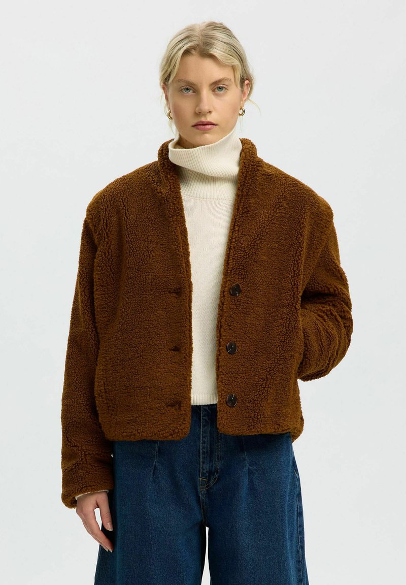 Femme portant une veste marron duveteuse boutonnée sur un pull à col roulé blanc et un jean bleu, debout avec une expression neutre.