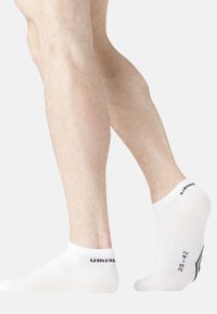Jambes portant des chaussettes blanches à coupe basse avec le texte noir "Umbro" et la taille de chaussure "39-42" visibles, sur un fond clair uni.