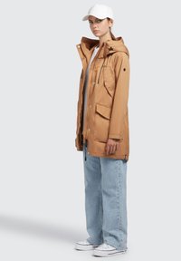 Hellbraune Kapuzenjacke mit Reißverschluss, mehreren Taschen und Kordelzug. Kombiniert mit hellblauen weiten Jeans und weißen Sneakern.