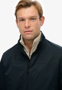 Chaqueta azul marino con cremallera sobre un suéter beis, con un cuello alto y textura suave, y con un detalle de cremallera en contraste.