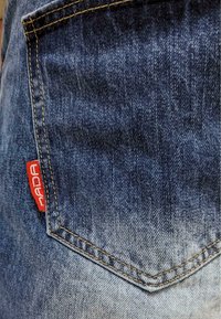 Denim fabric with a gradient from dark to light blue. Features a visible back pocket with a red branded label and yellow stitching accents.