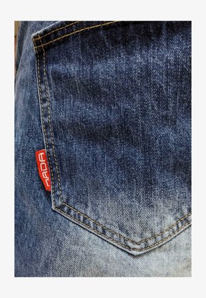 Tela de mezclilla con un degradado de azul oscuro a azul claro. Presenta un bolsillo trasero visible con una etiqueta de marca roja y detalles de costura amarilla.