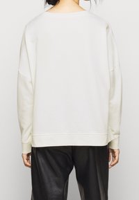 Bílá oversize mikina s nízkými rameny, žebernatými manžetami a kulatým výstřihem, spojená s černými kalhotami z umělé kůže.