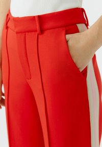 Rode op maat gemaakte broek met plooien en een zijzak, met een contrasterende verticale streep in beige en lichtroze.