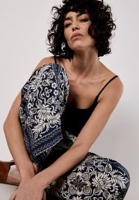 Hosen mit Marine-Muster und cremefarbenen Blumenmotiven; weit geschnitten und aus weichem Stoff. Schwarzes Tanktop, braune Sandalen. Modell sitzt mit entspannter Haltung.