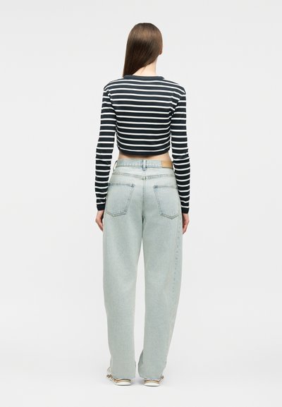 Γυναίκα που στέκεται με την πλάτη γυρισμένη, φοράει μακρυμάνικη κορυφή κοντού μήκους με ασπρόμαυρες ρίγες, φαρδιά ανοιχτό μπλε τζιν και αθλητικά παπούτσια σε ουδέτερο φόντο.