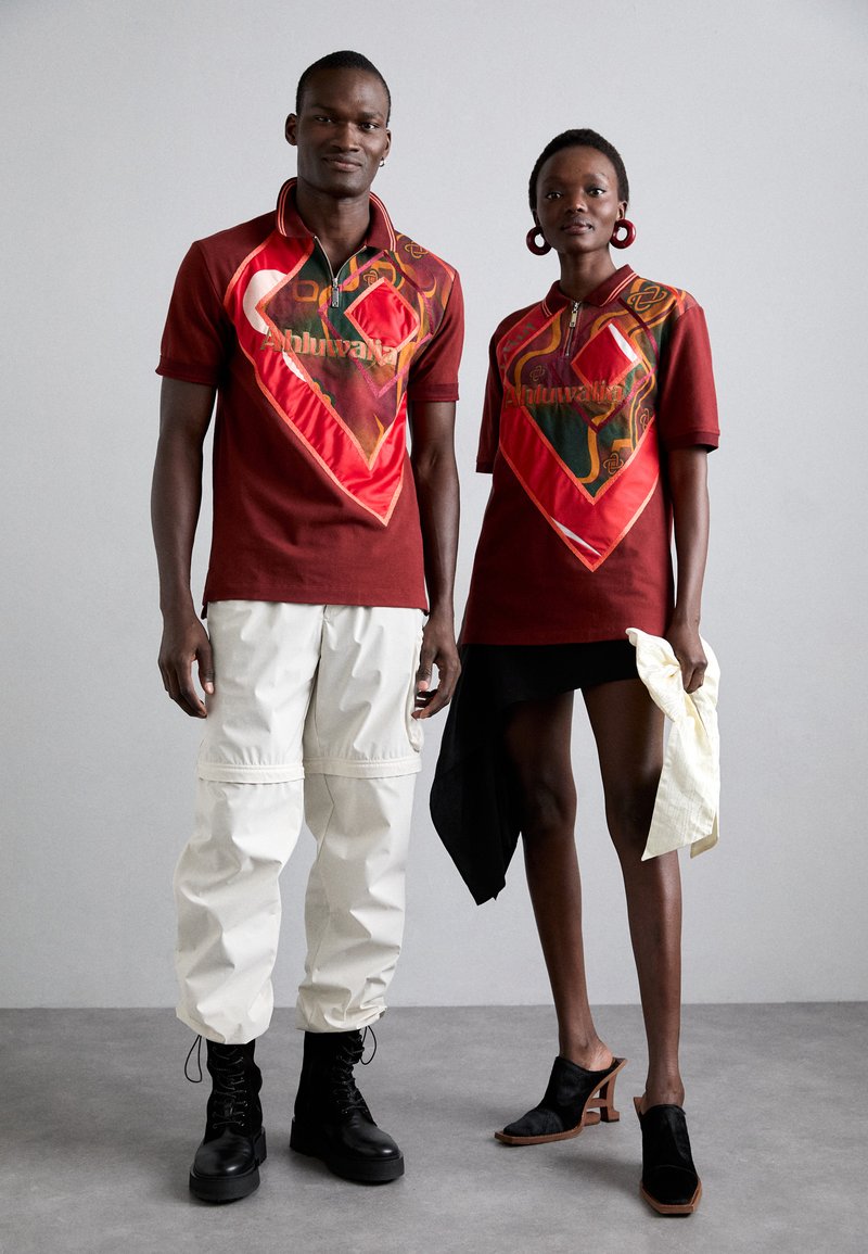 Een paar modellen in bijpassende rode polo shirts met grafische ontwerpen. Het ene model draagt beige cargobroeken; het andere model draagt een zwarte asymmetrische rok en heeled mules.