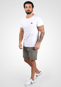 T-shirt blanche à manches courtes avec un logo triangulaire noir, associée à un short texturé gris et des baskets blanches avec des accents rouges et bleus.