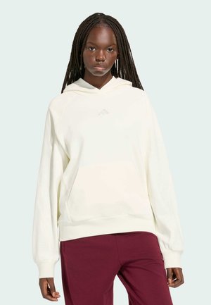 Junge Frau mit langen Zöpfen, die einen übergroßen weißen Hoodie und bordeauxrote Hose trägt, steht vor einem schlichten hellen Hintergrund.