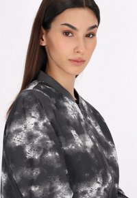 Jachetă bomber neagră cu un model alb asemănător norilor, guler ribijat și fermoar frontal. Materialul pare neted și ușor lucios.