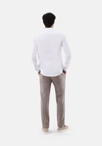 Camicia bianca a maniche lunghe con colletto standard, abbinata a pantaloni a quadri marrone chiaro e sneaker beige, vista da dietro.
