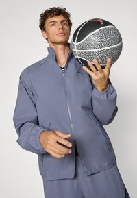 Szara sportowa kurtka z wysokim kołnierzem, pełnym zamkiem błyskawicznym i siateczkowymi detalami. Połączona z czarno-szarym teksturowanym koszem do koszykówki.