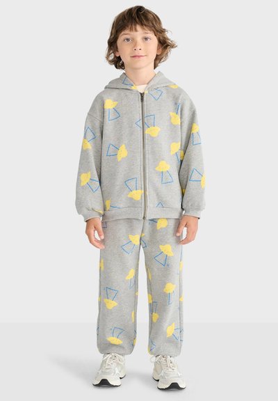 Enfant portant un sweat à capuche gris et un pantalon avec des motifs de poussin jaune et de triangles bleus, debout et face à l'avant sur un fond blanc.