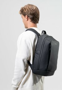Mochila negra y estructurada con correas acolchadas. Fabricada con material suave, presenta un zipper en la parte superior y bolsillos laterales para almacenamiento adicional.