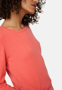 Pull en corail avec un col rond, fabriqué à partir d'un tissu doux. Présente des manches longues et une silhouette ajustée, mettant en avant une texture lisse.