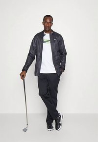 Črno vetrovko preko bele majice Nike z zelenim logotipom, kombinirano s črnimi hlačami in čevlji. Drži golf palico.