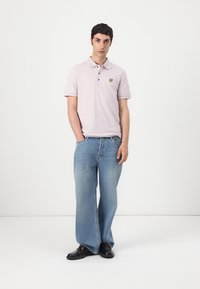 Polo shirt violette claire avec deux boutons et un logo jaune sur la poitrine, associé à un jean bleu à jambes larges et des chaussures noires.