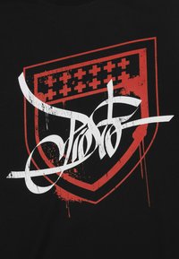 Czarny t-shirt z dużą czerwoną grafiką tarczy oraz białym tekstem w stylu graffiti, z plamami i przetartymi detalami.