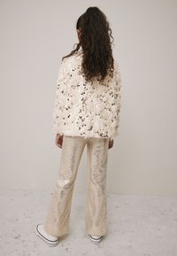 White fluffy cardigan with sequins paired with beige flared pants featuring a shiny, reflective texture. Model wearing white platform sneakers.