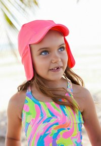 Cappello da sole rosa con alette per le orecchie estese, realizzato in materiale leggero. Indossato con un costume da bagno colorato e fantasioso con vivaci vortici.