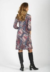 Kobieta z długimi falowanymi włosami, ubrana w wzorzystą sukienkę do kolan oraz czarne, wysokie kozaki na obcasie, stojąca tyłem na białym tle.