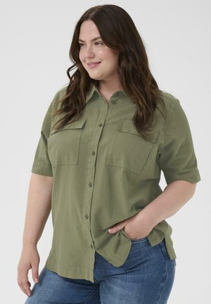 Uśmiechnięta kobieta o długich brązowych włosach, ubrana w oliwkową koszulę na guziki z krótkim rękawem i niebieskie dżinsy, z jedną ręką w kieszeni.