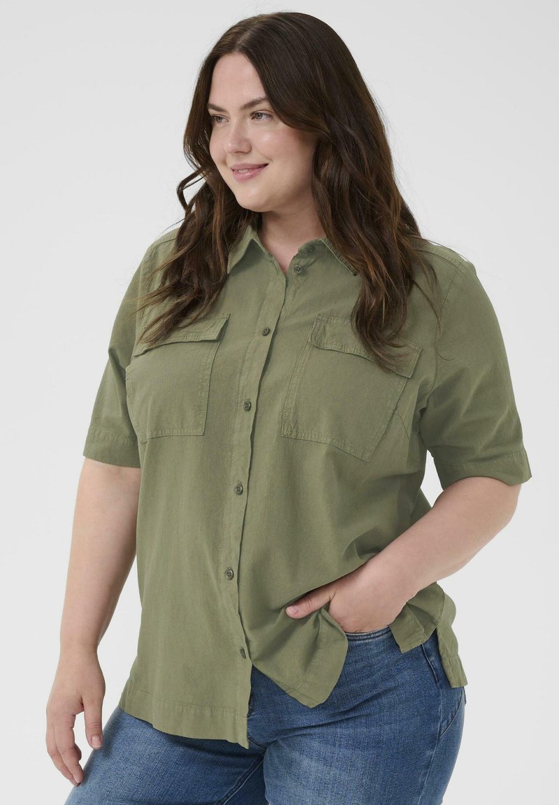 Femme souriante aux longs cheveux bruns portant une chemise courte à manches courtes kaki boutonnée et un jean bleu, une main dans la poche.