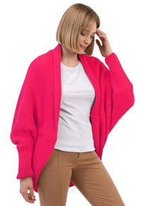 Cárdigan de punto rosa intenso con mangas anchas, textura acanalada y un frente abierto, llevado sobre una camisa blanca. Pantalones ajustados color beige completan el look.