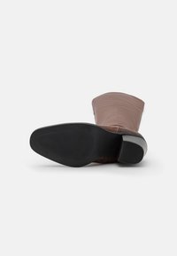 Bota de cuero marrón hasta la rodilla con punta redondeada, acentos texturizados, un tacón bloque moderado y una suela de goma flexible.