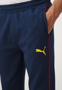 Mornarsko modri trenirki z elastičnim pasom, ki imata rdeče stranske črte in rumeni puma logotip spredaj. Mehka tekstura tkanine.