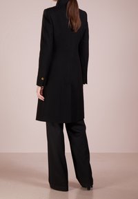 Cappotto di lana nera con vestibilità su misura, colletto alto e dettagli in bottone dorato sulle maniche, abbinato a pantaloni neri a gamba larga.