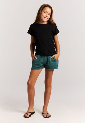 Juodi trumpomis rankovėmis marškinėliai derinami su žalsvomis šortais. Šortai turi elastingą juosmenį, šonines kišenes ir sukarpytą apvadą. Juodos šlepetės.