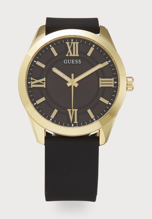 Montre dorée avec un bracelet en silicone noir, cadran rond, affichage noir et index en chiffres romains. Présente des aiguilles argentées et un logo de la marque.