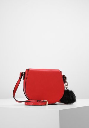 Rød skuldertaske med justerbar rem og sort fluffy pompom-nøglering på en hvid piedestal mod en ensfarvet baggrund.