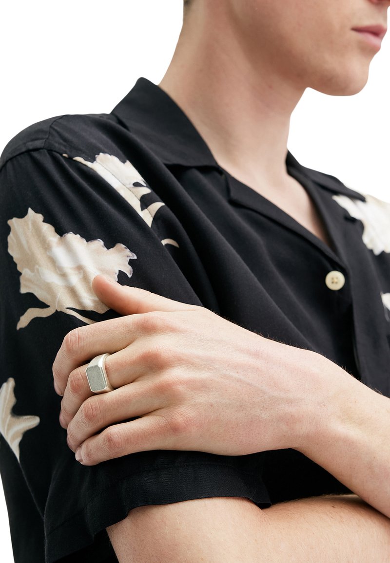 Black shirt with cream floral prints, short sleeves, and button closure. Silver hexagonal ring on hand resting on arm. Smooth texture.