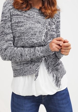 Frau trägt eine graue, gestrickte, mit Fransen versehene Strickjacke über einer weißen Bluse, kombiniert mit blauen Jeans, die Hände vor dem Körper verschränkt.