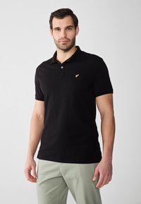 Črna polo majica iz bombaža, z ovratnikom, kratkimi rokavi in majhnim rumenim logotipom na prsih. Gladek, klasičen dizajn.