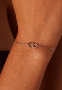 Zilveren kettingarmband met twee verstrengelde hartvormen, waarvan er één glad is en de ander is versierd met kleine kristallen.