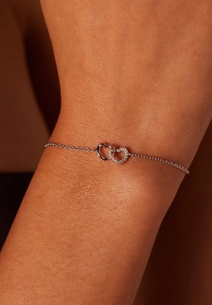 Zilveren kettingarmband met twee verstrengelde hartvormen, waarvan er één glad is en de ander is versierd met kleine kristallen.