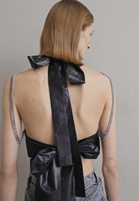 Black satin backless top with a large bow and chain accents on straps. Smooth texture, clean lines, and a minimalist design.