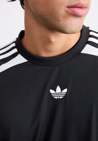 Musta urheilupaita pystysuorilla raidoilla, pyöreä pääntie, valkoinen Adidas-logo ja kolme valkoista olkaviivaa, sileä tekstuuri.