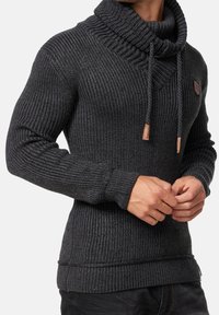 Pull en tricot gris foncé avec un col haut, présentant une texture côtelée, des cordons de serrage et une petite étiquette en cuir de marque sur la poitrine.