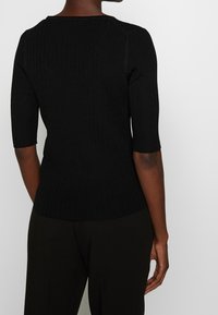 Musta ribattoppi, jossa on kolme neljäsosaa hihoja ja kireä muotoilu. Pehmeä tekstuuri ja pyöreä pääntie, mikä korostaa virtaviivaista siluettia.